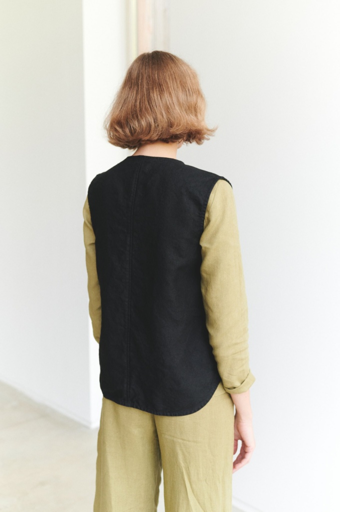 Back of a model in a black linen vest worn over a green long sleeve linen top
