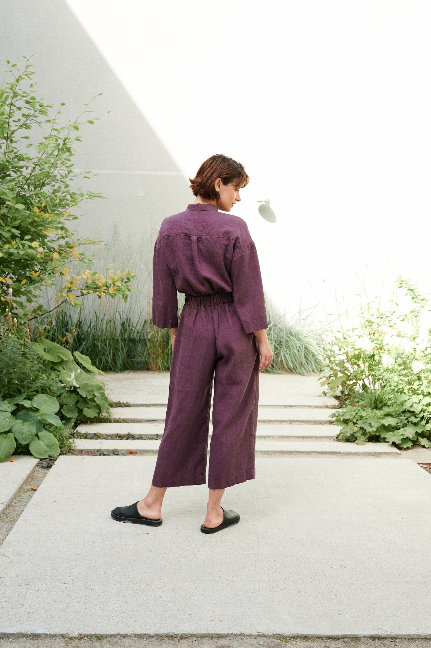 Back of a model in a violet linen jumpsuit with wide, lightly cropped legs