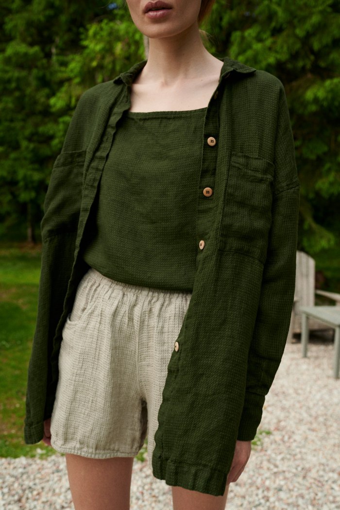 An oversized green waffle linen button down shirt with wooden buttons worn over a linen summer top and shorts