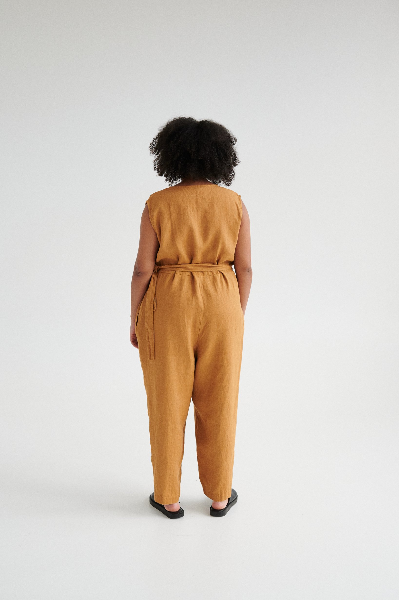 Back of a model in an oversized sleeveless linen jumpsuit with a belt tied around the waist