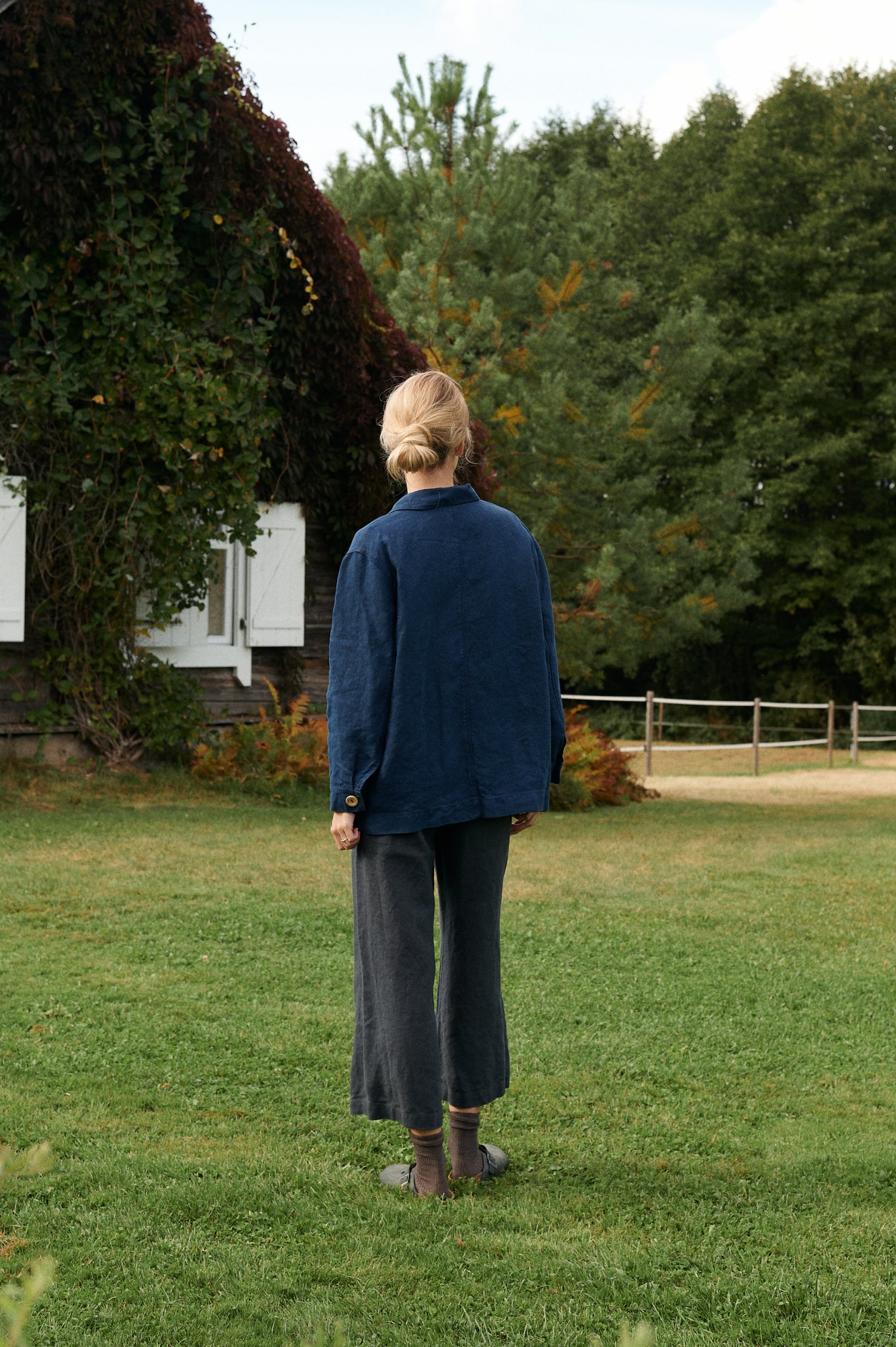 Back of a navy blue heavy linen utility jacket paired with cropped grey linen trousers