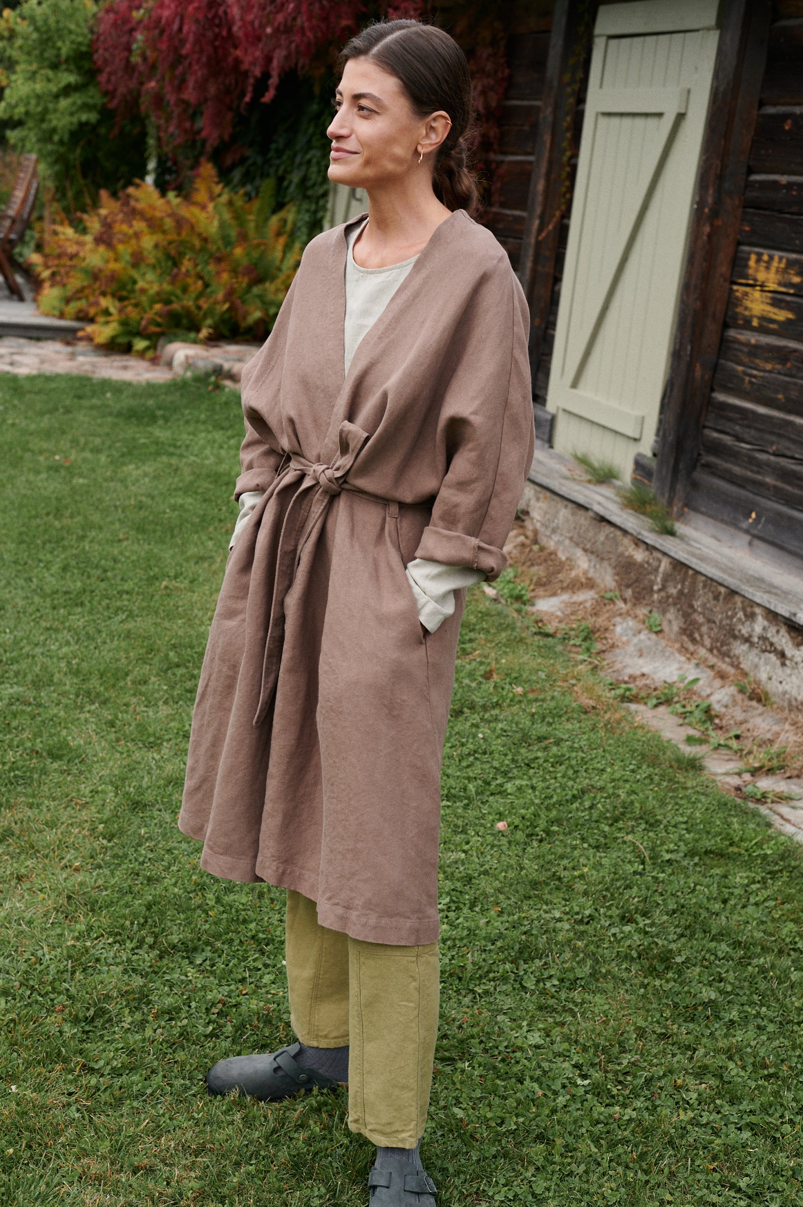 A woman wearing a long heavy cacao linen jacket with slightly rolled up loose sleeves and hands in pockets