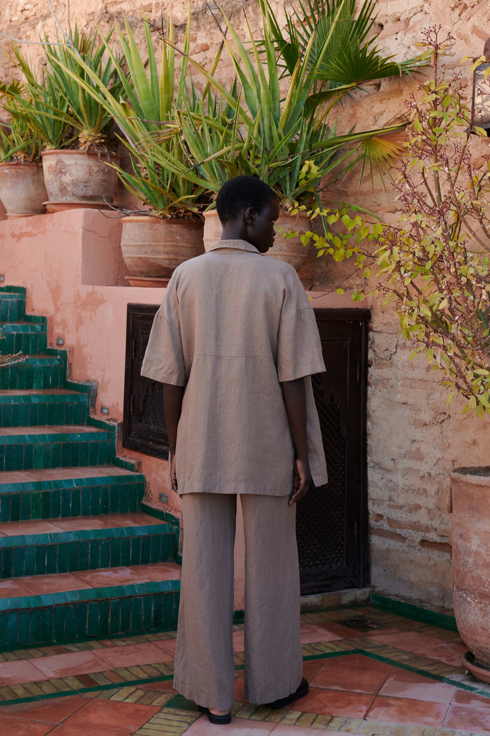 a back view of a model wearing a linen set of short-sleeved linen shirt, and wide leg trousers in mocha mousse color