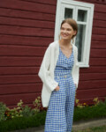 model smiling wearing blue gingham linen set of relaxed trousers and wrap top with an oversized linen jacket in milky white