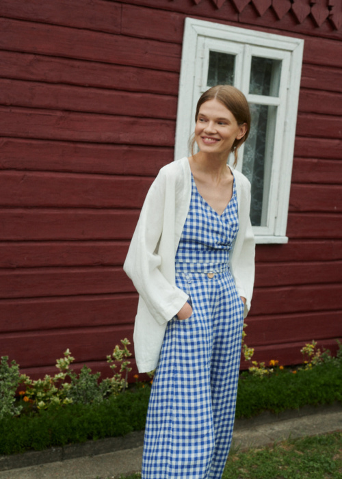 model smiling wearing blue gingham linen set of relaxed trousers and wrap top with an oversized linen jacket in milky white