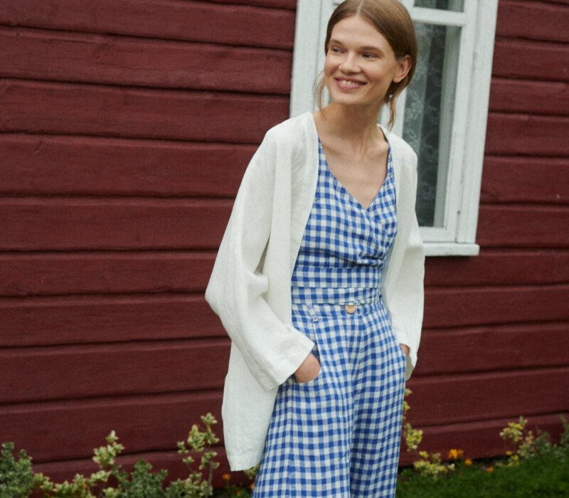 model smiling wearing blue gingham linen set of relaxed trousers and wrap top with an oversized linen jacket in milky white