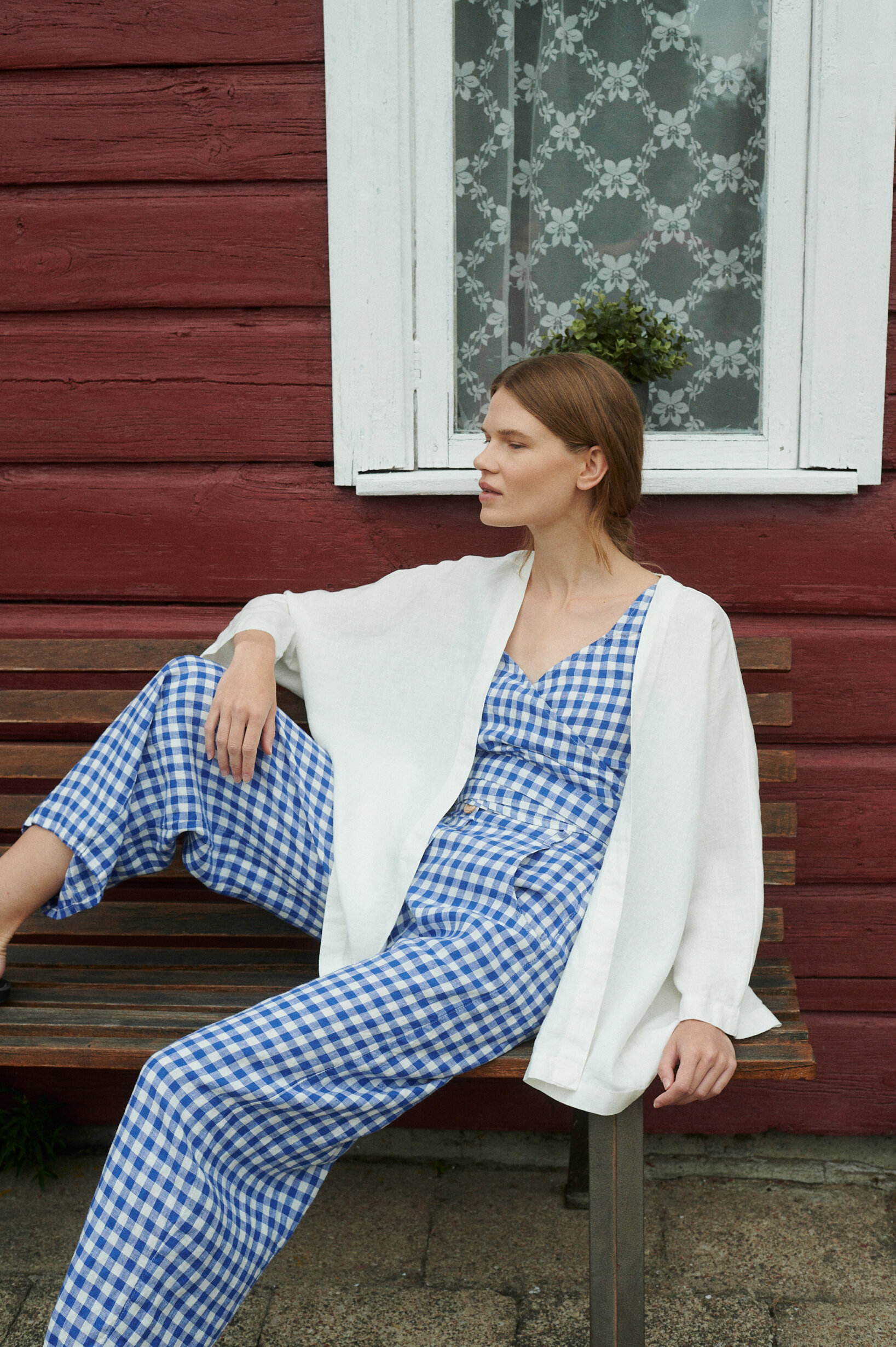 model sitting near the summer cottage wearing blue gingham linen set of relaxed barrel trousers and wrap top with an oversized linen jacket in milky white