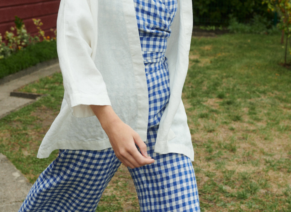 model on a move wearing blue gingham linen set of relaxed trousers and wrap top with an oversized linen jacket in milky white