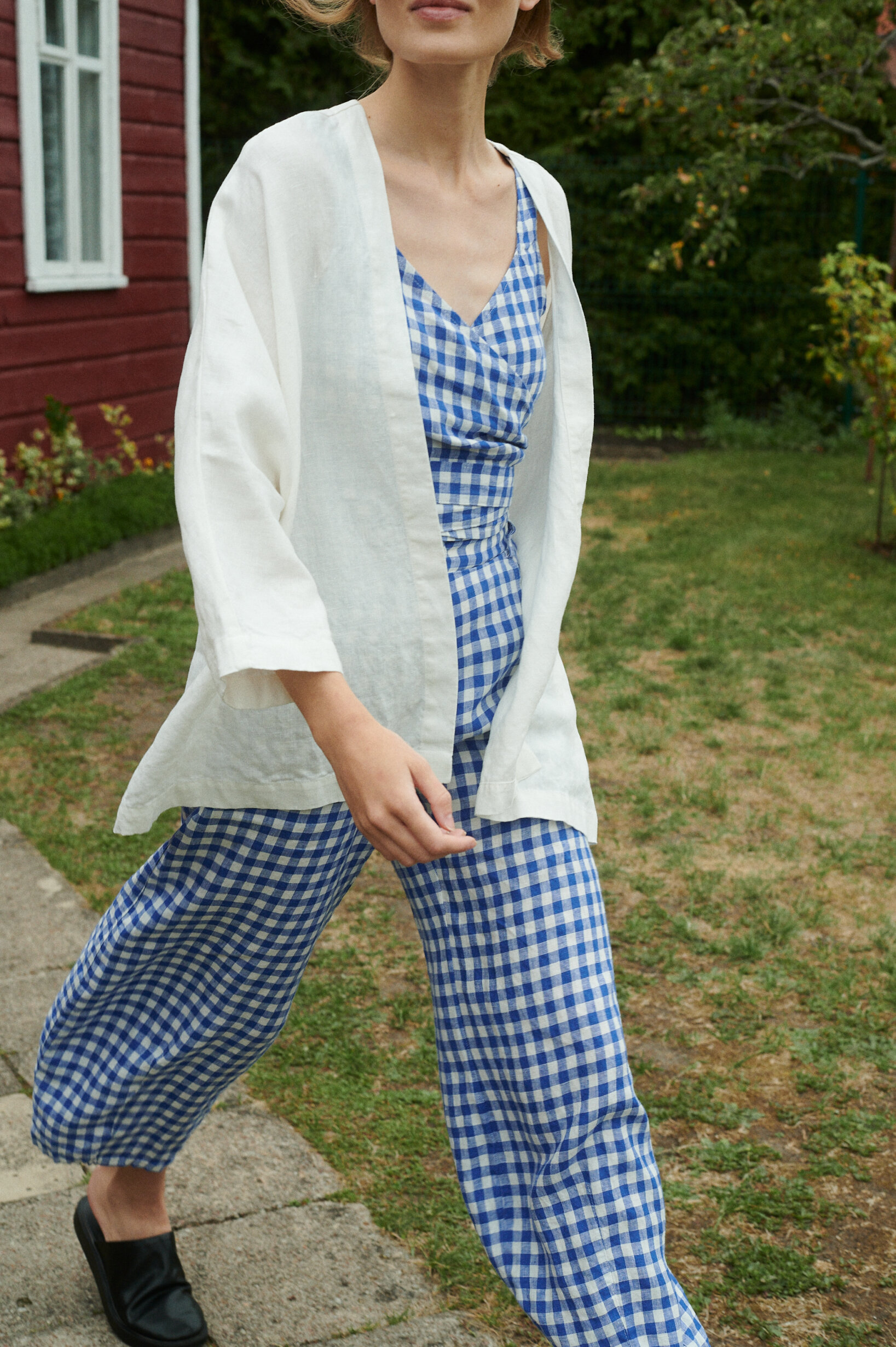 model walking and wearing blue gingham linen set of relaxed trousers and wrap top with an oversized linen jacket in milky white