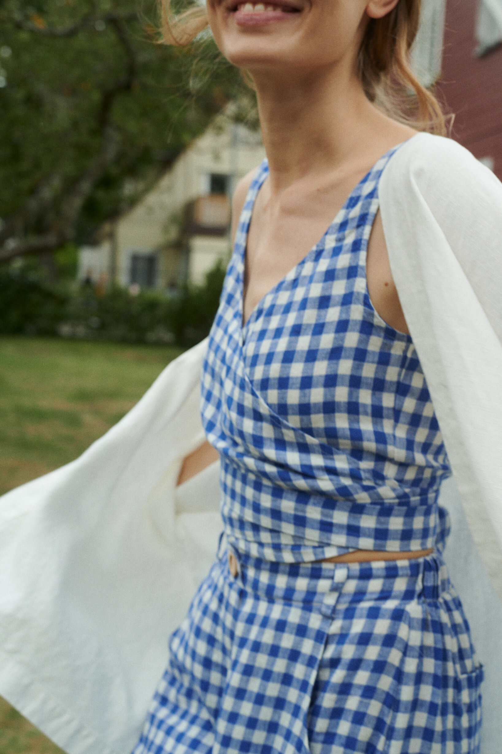 a close up of model wearing blue gingham summer linen set of relaxed trousers and wrap top with an oversized linen jacket in milky white