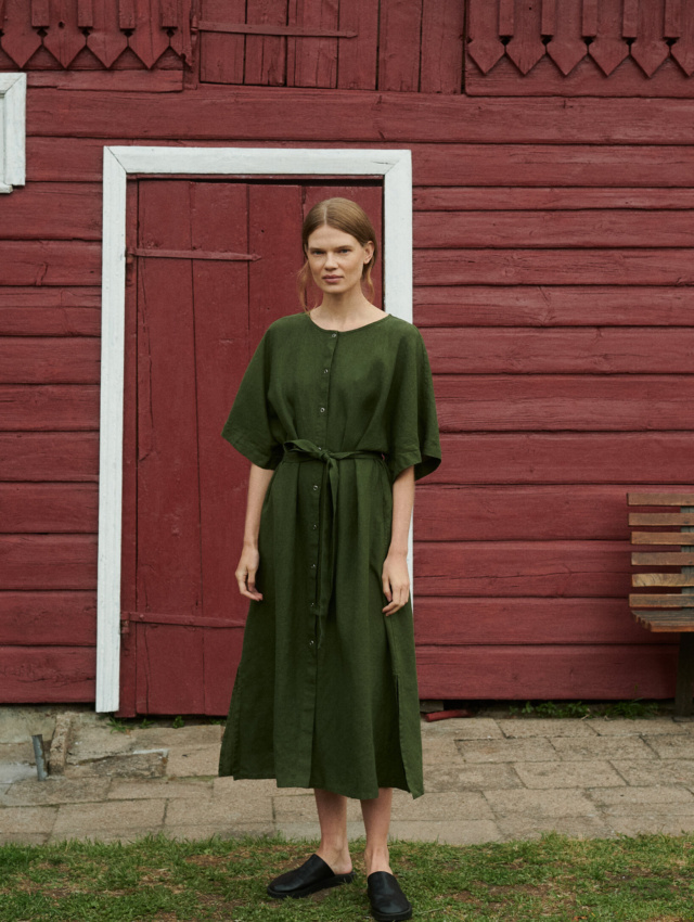 model posing outside the red summer house in an oversized linen dress in forest green with snap openings and belt