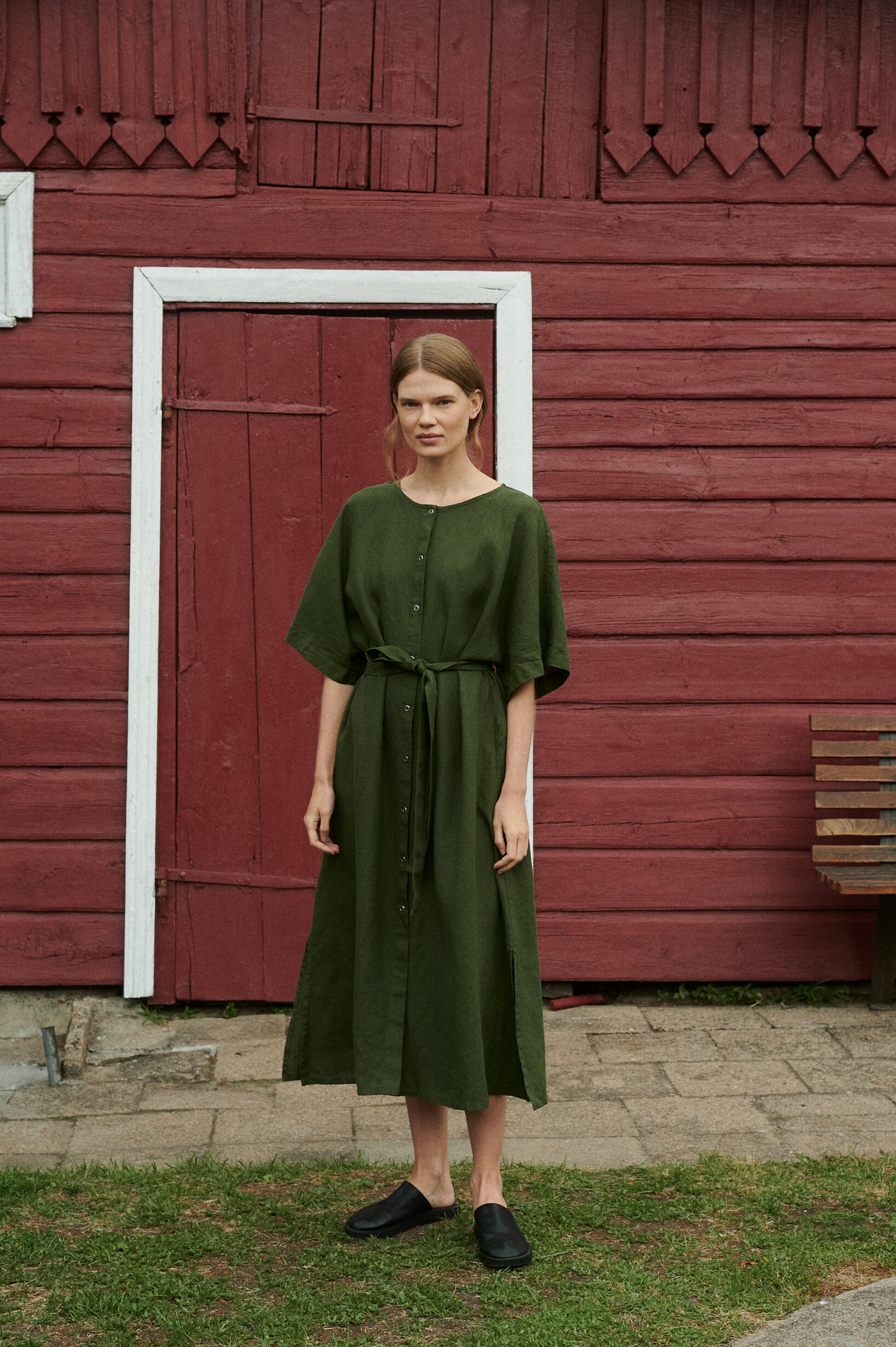 model posing outside the red summer house in an oversized linen dress in forest green with snap openings and belt