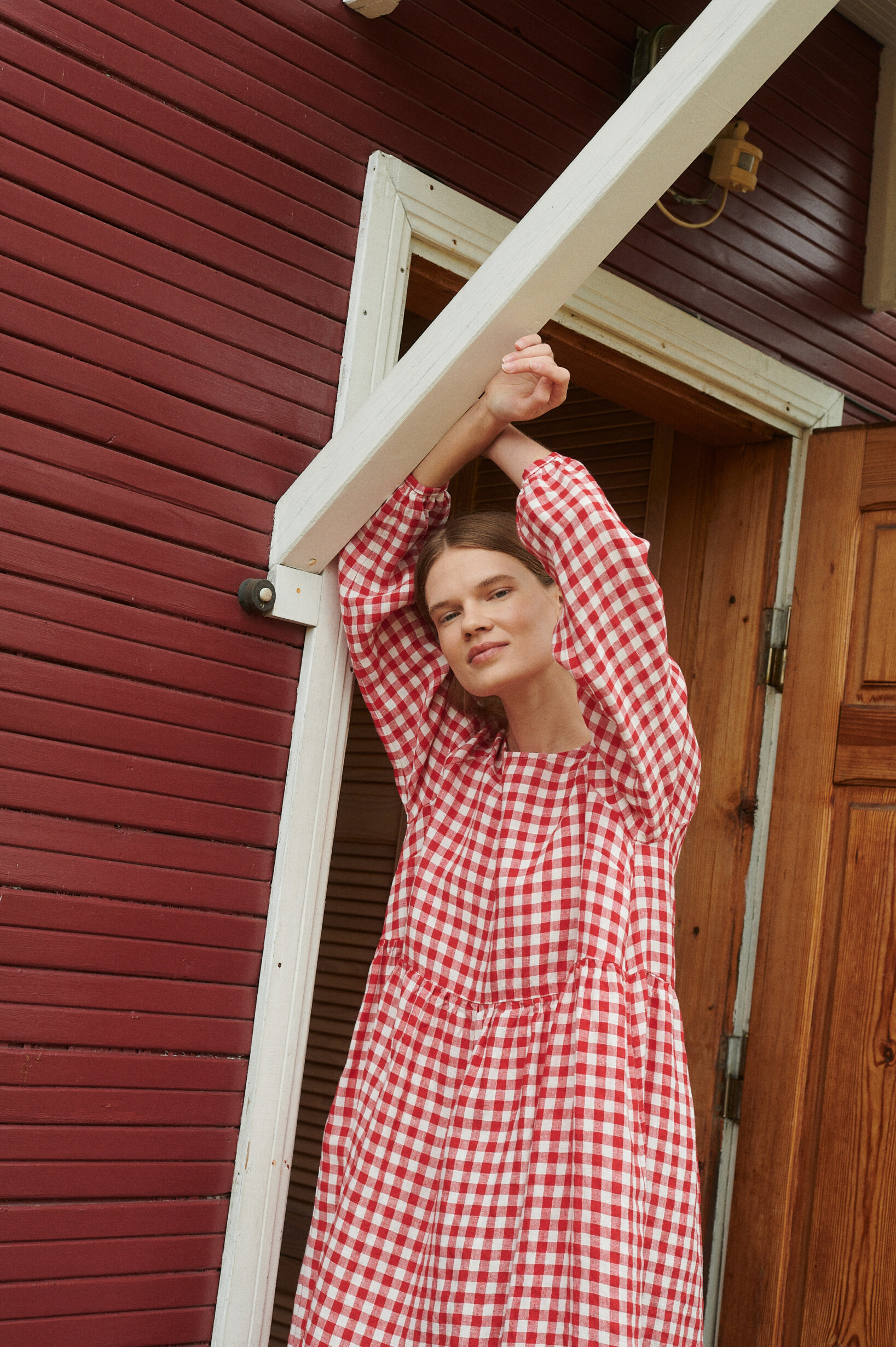 model leaning on a porch wearing red gingham oversized linen dress with full length sleeves