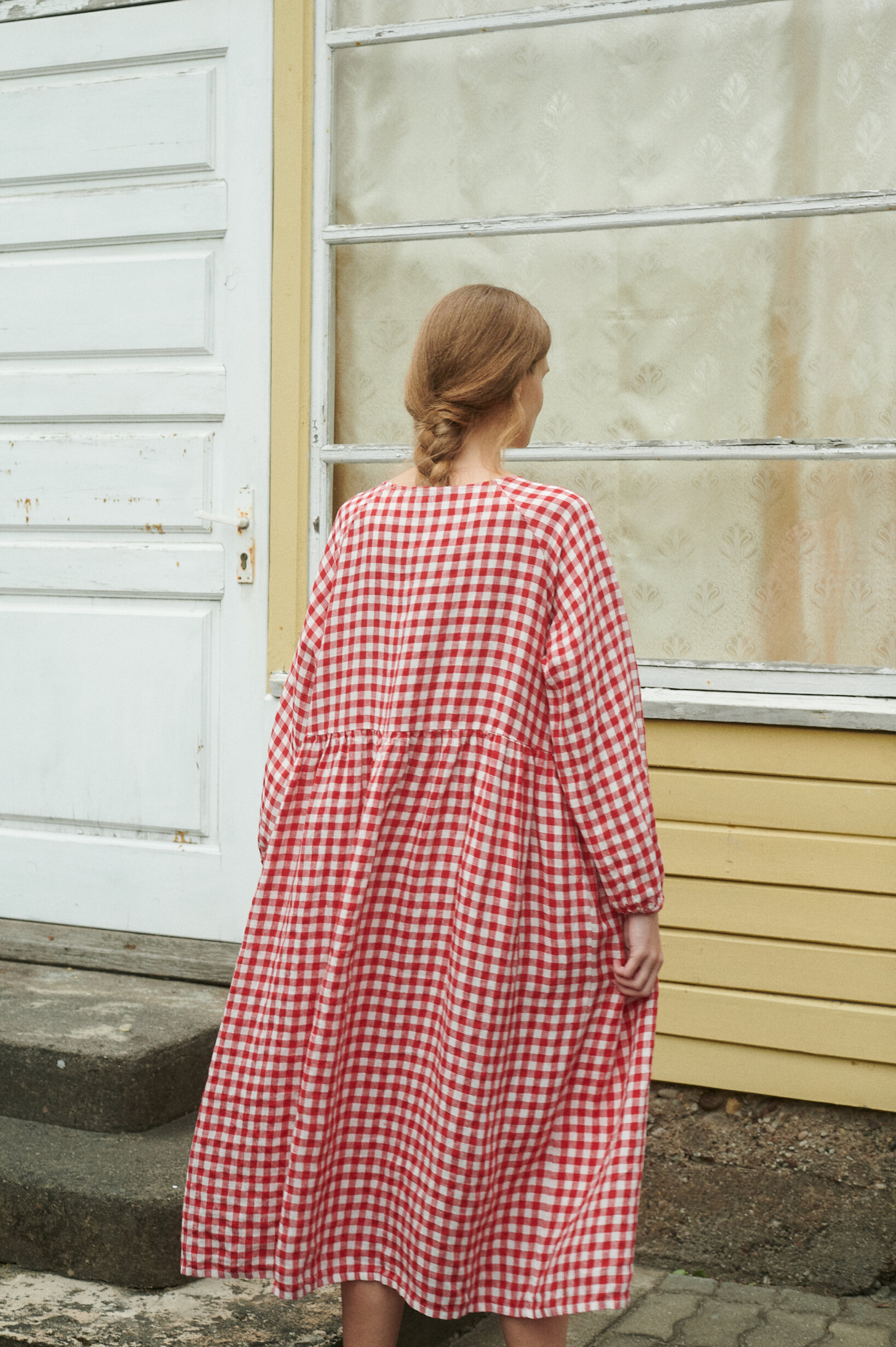 a back view of a model wearing oversized red gingham linen dress near the summer house