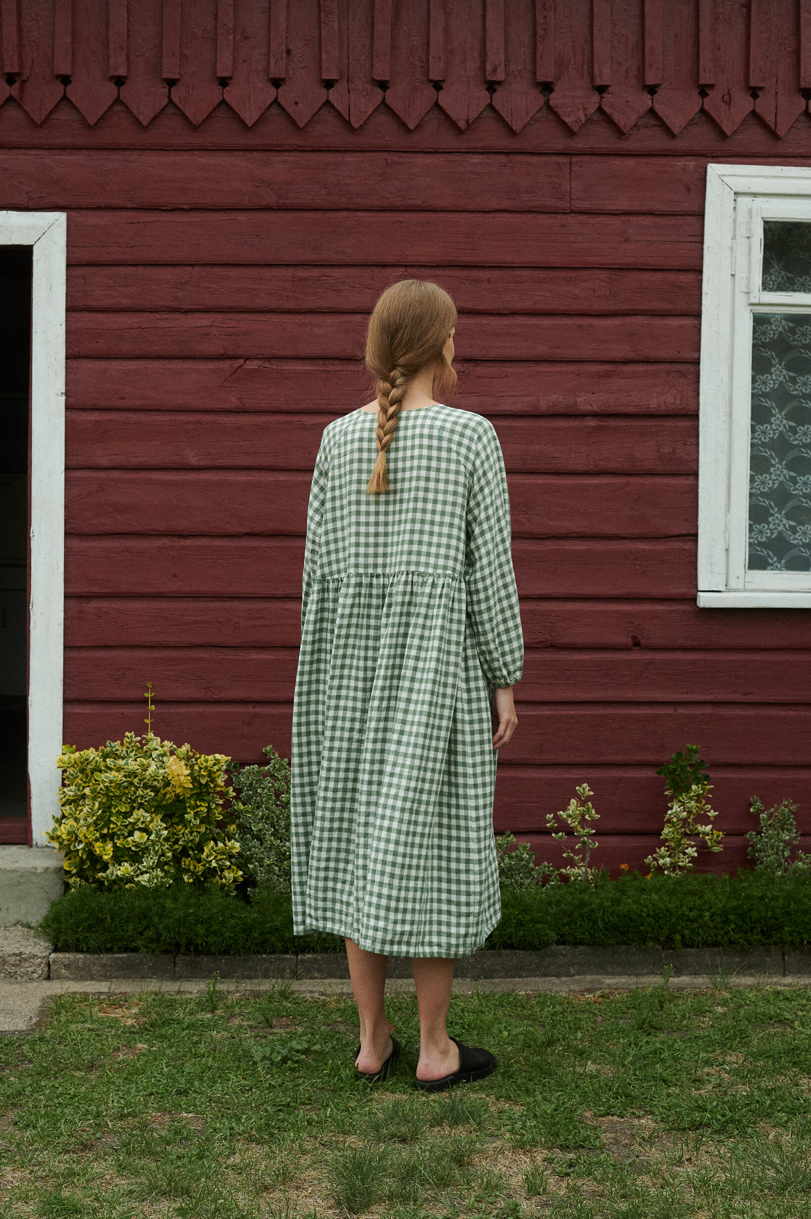a back view of model in a garden wearing oversized green gingham linen dress with full length sleeves and pockets