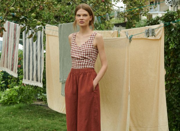 a woman standing near the drying sheets outside the garden wearing wide leg trousers in terracotta linen that is matched with V-neck wrap top in terracotta gingham pattern