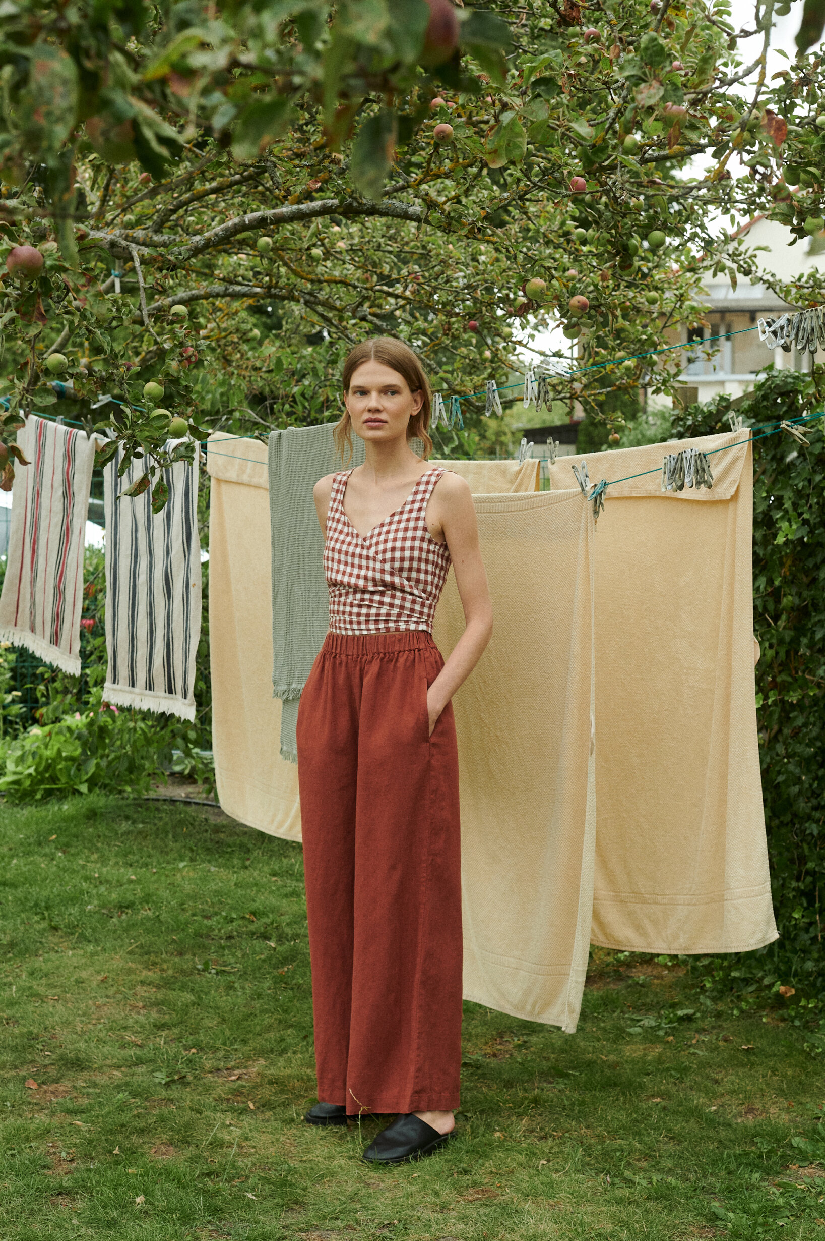 a woman standing near the drying sheets outside the garden wearing wide leg trousers in terracotta linen that is matched with V-neck wrap top in terracotta gingham pattern