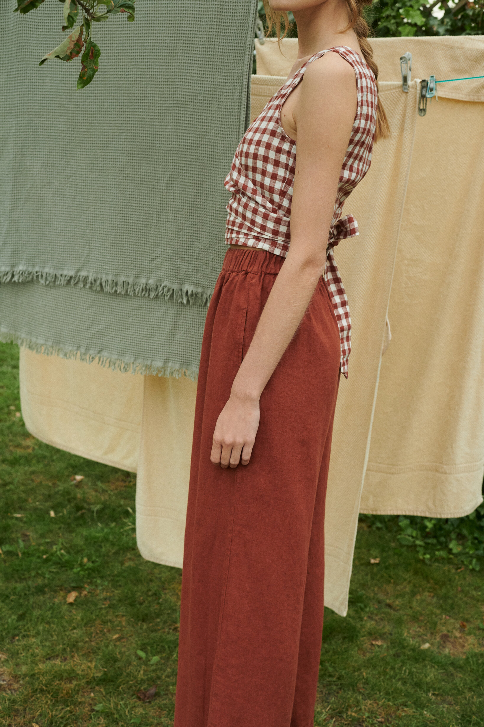 a close up of a model wearing a terrracotta gingham linen wrap top and wide leg trousers in terracotta linen near the sheets drying outside