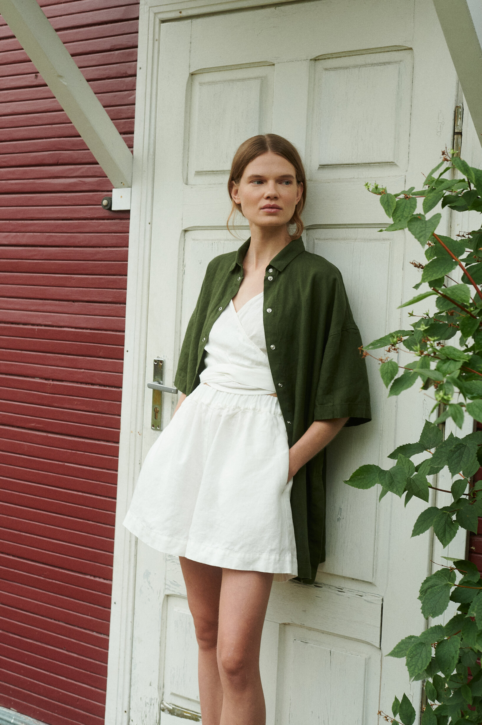 a model in a garden wearing short sleeved linen shirt in forest green matched with wrap top and wide shorts in milky white linen
