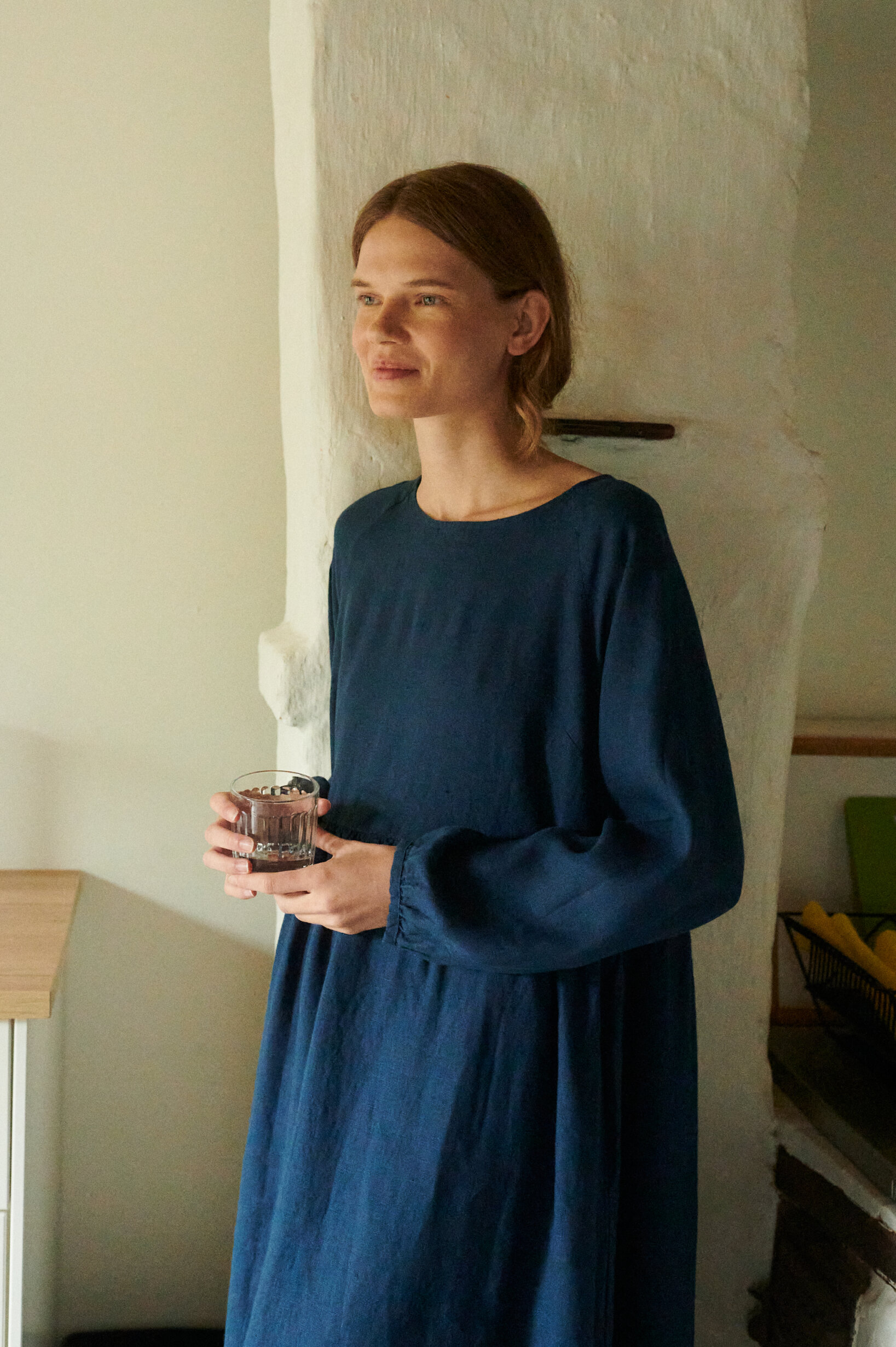 a close up of model wearing oversized linen dress with full length sleeves in navy blue holding a glass of water