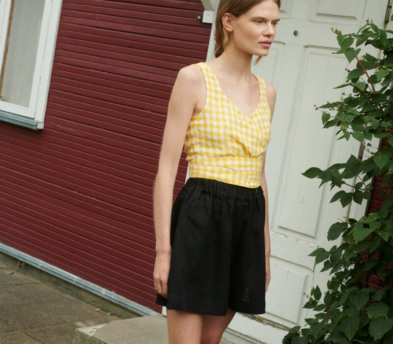 a girl wearing linen wrap top in yellow gingham and black linen shorts near the summer house