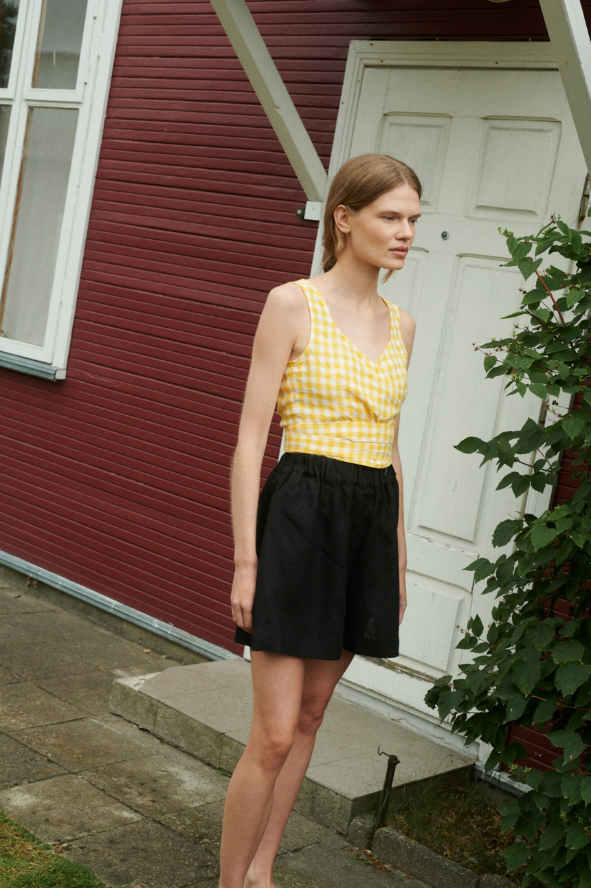 a girl wearing linen wrap top in yellow gingham and black linen shorts near the summer house