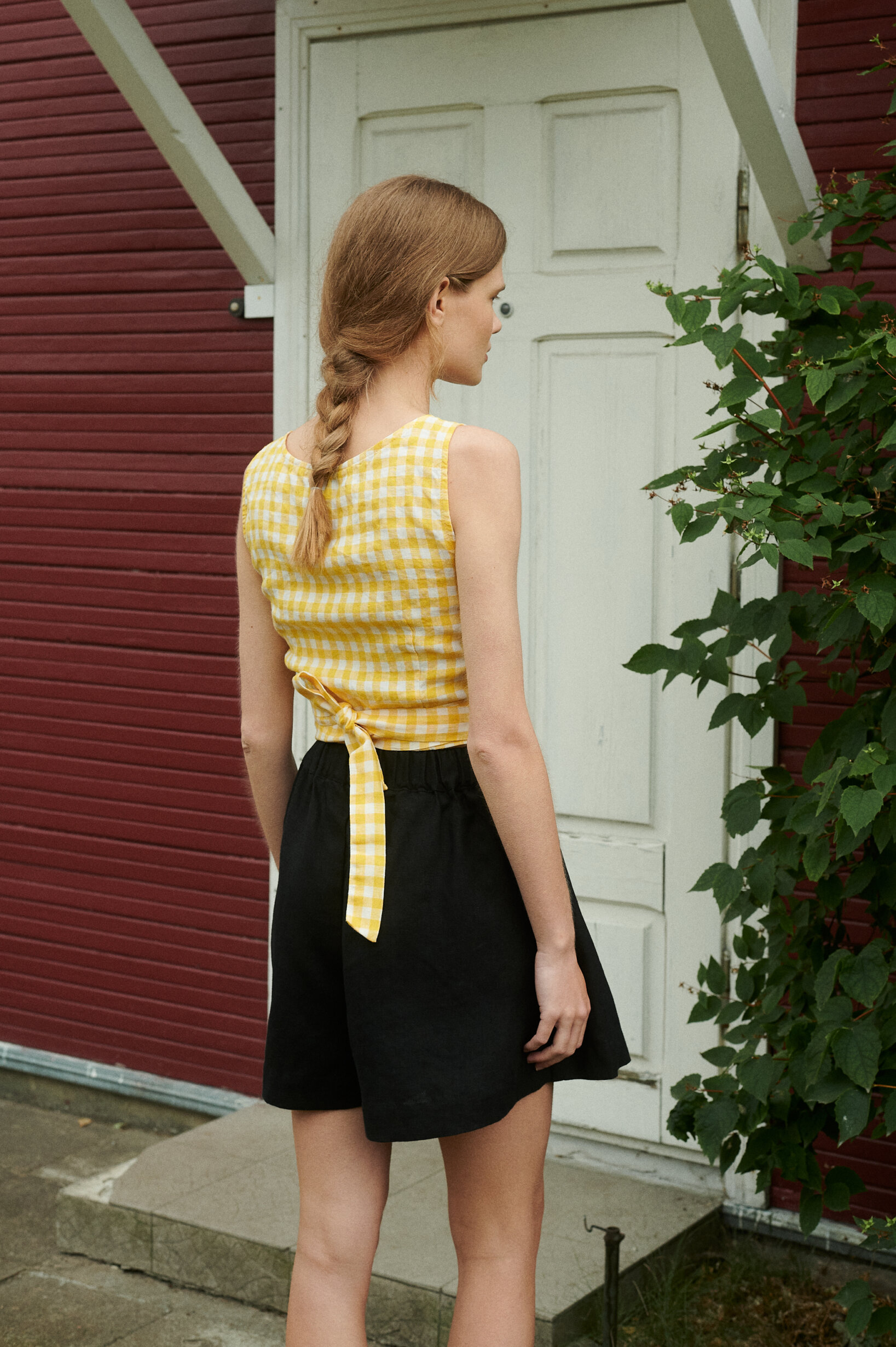 the back view of a model standing outside the red house wearing yellow gingham linen wrap top and black linen shorts