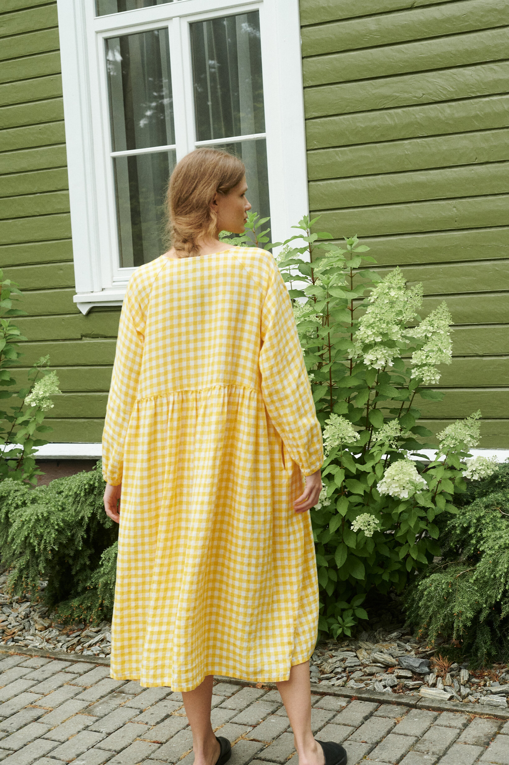 a back view of a model in a garden wearing a yellow gingham linen dress in oversized fit and full length sleeves