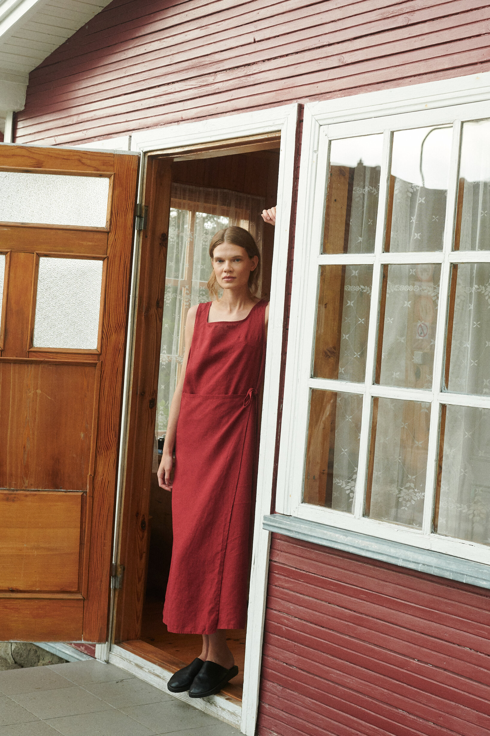 model posing with a full length sleeveless summer wrap dress in burgundy red linen in a summer cottage