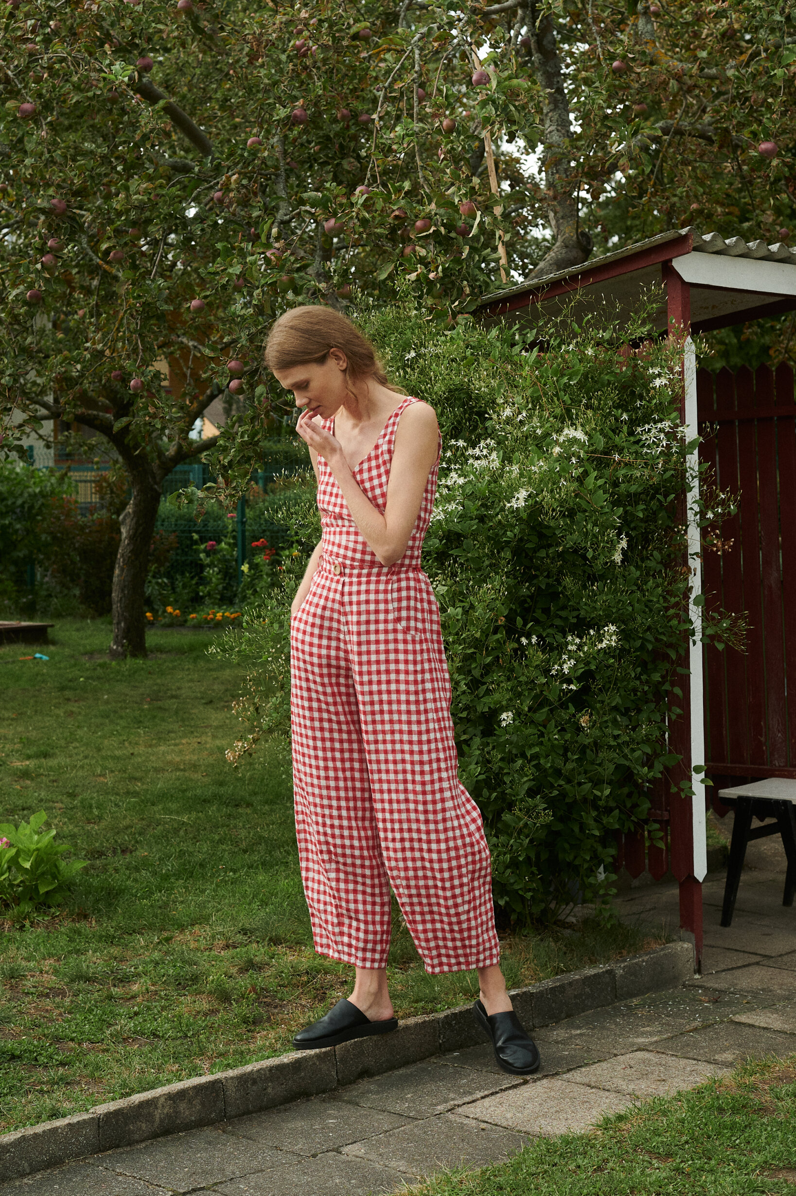 Model in the garden wearing summer linen set of wrap top and relaxed barrel leg trousers in red gingham linen.