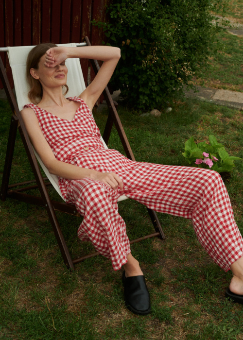 Model sitting on a chair wearing summer linen set of V-neck wrap top and barrel leg trousers in red gingham linen.