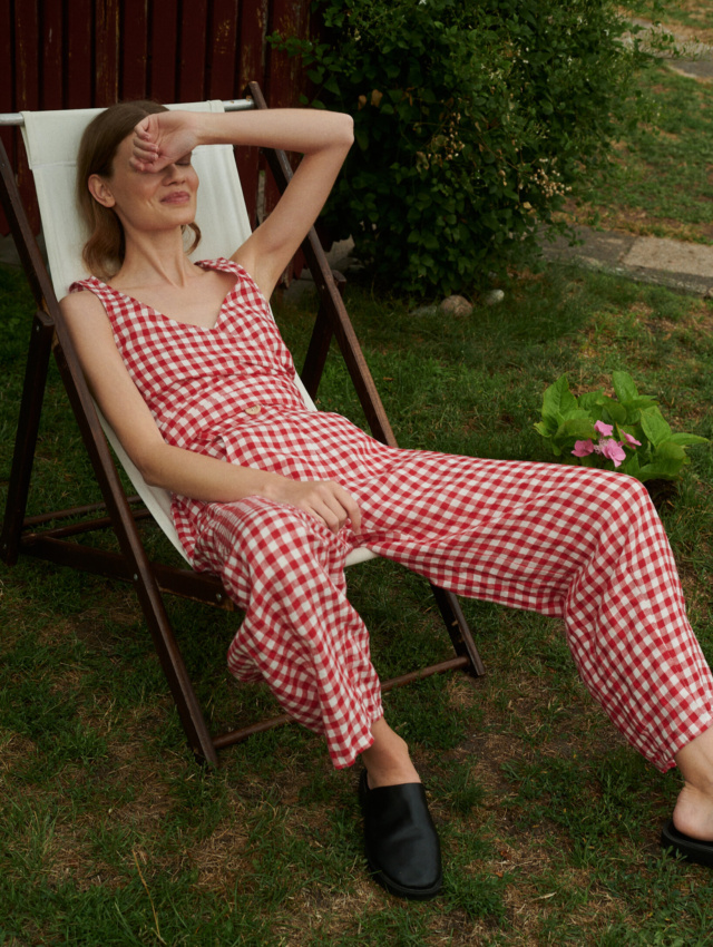 Model sitting on a chair wearing summer linen set of V-neck wrap top and barrel leg trousers in red gingham linen.