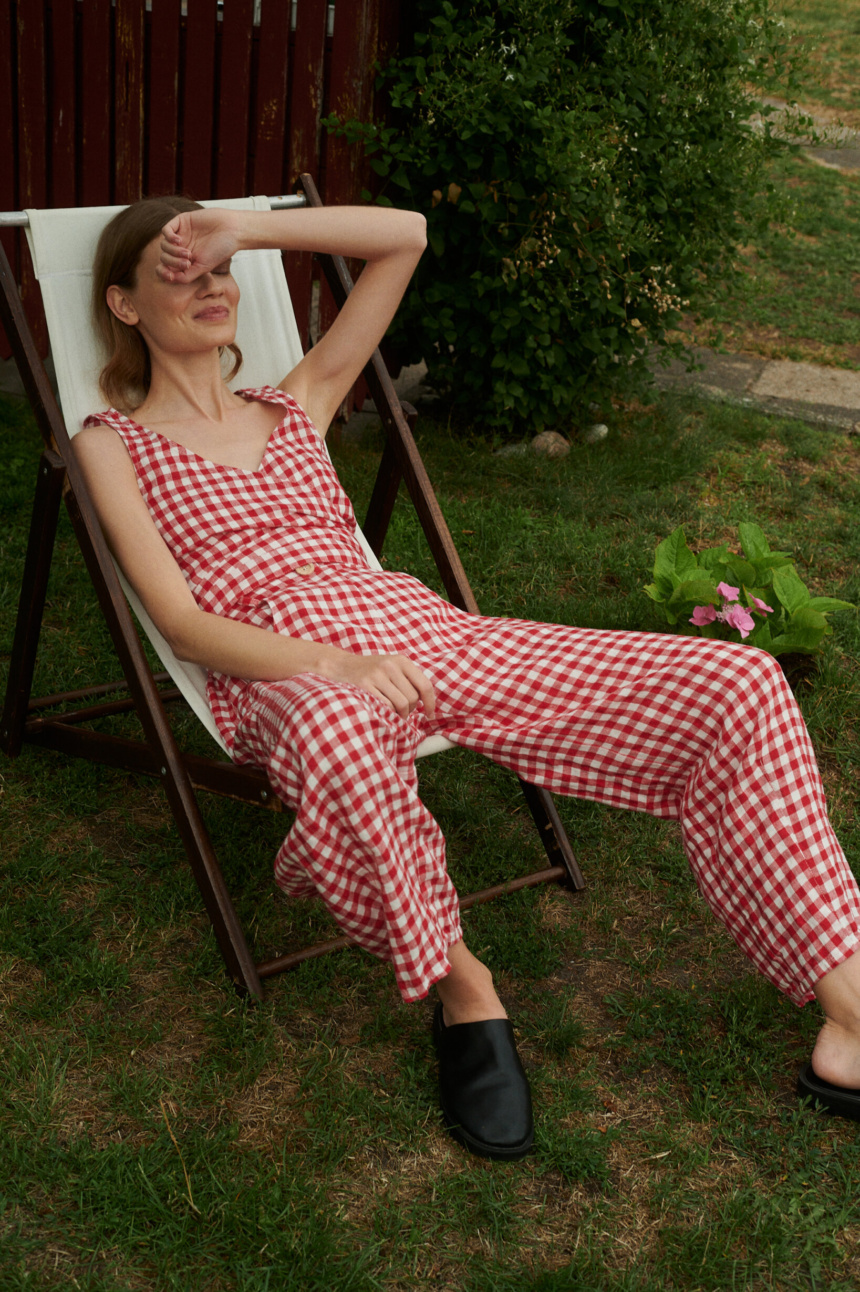 Model sitting on a chair wearing summer linen set of V-neck wrap top and barrel leg trousers in red gingham linen.