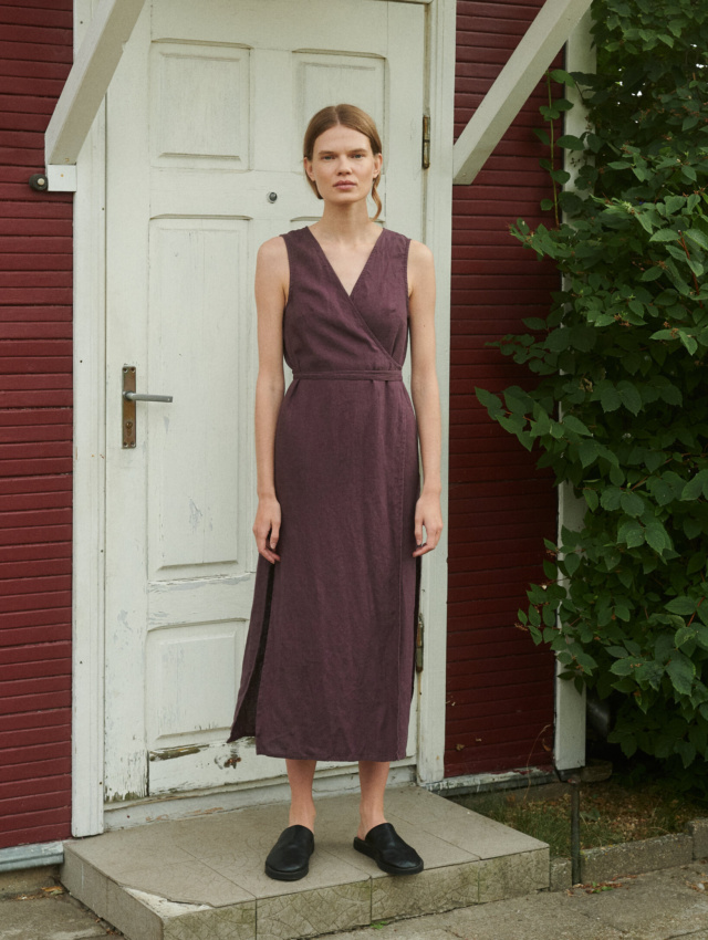 model wearing a V-neck linen wrap dress in eggplant violet with side slits and midi length while standing outside summer house