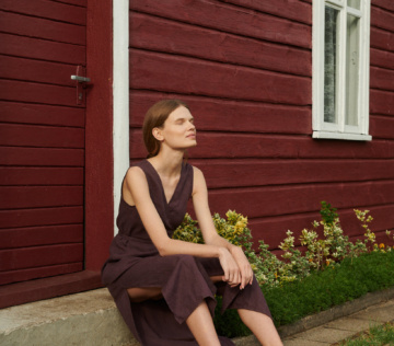 a woman posing while sitting down wearing a V-neck linen wrap dress in eggplant violet with side slits near the red summer cottage