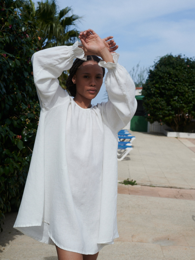 Woman posing in mini length oversized linen dress with full-length balloon sleeves end with elasticated ruffled cuffs. In a milky white linen color.