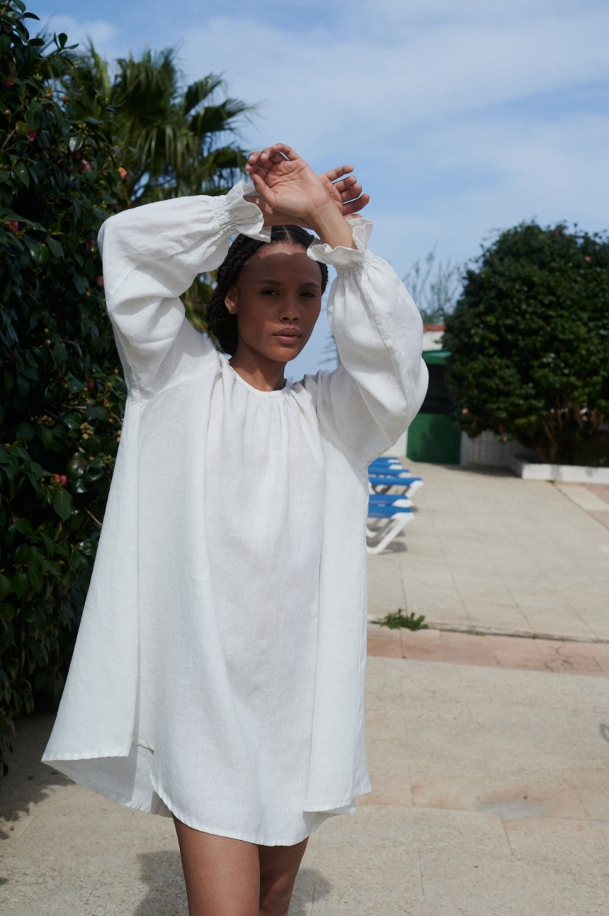 Woman posing in mini length oversized linen dress with full-length balloon sleeves end with elasticated ruffled cuffs. In a milky white linen color.