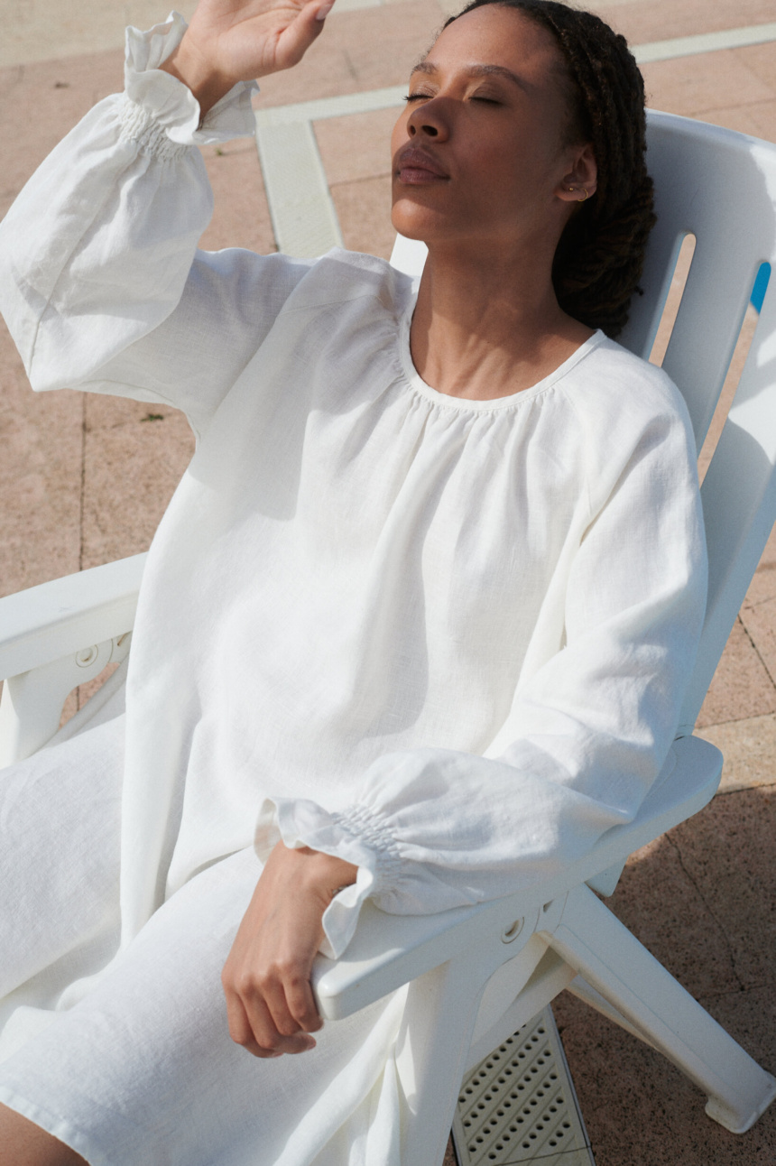 woman sitting in a milky white linen Agata dress with a mini-length hem, oversized fit, and full-length balloon sleeves