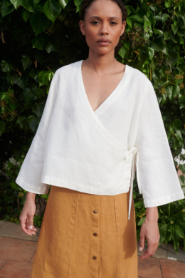 Model wearing a loose-fitting white linen top with a deep V-neck and long kimono sleeves paired with a camel linen skirt with metal snap buttons at the front