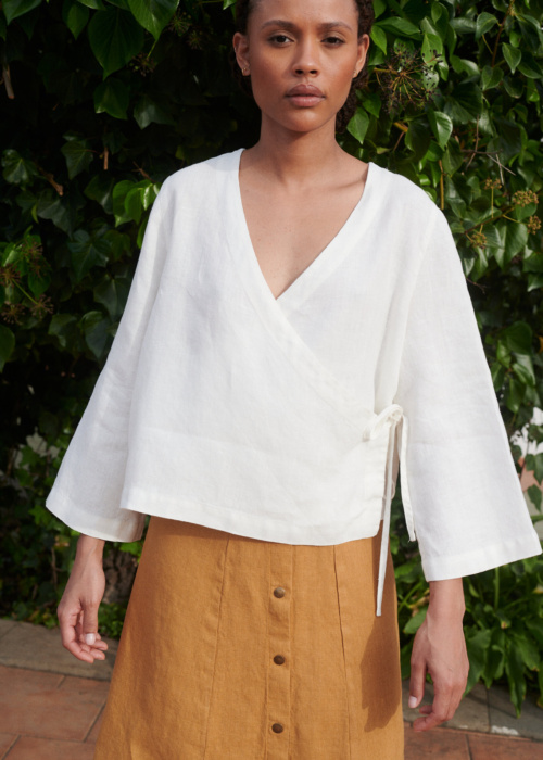 Model wearing a loose-fitting white linen top with a deep V-neck and long kimono sleeves paired with a camel linen skirt with metal snap buttons at the front