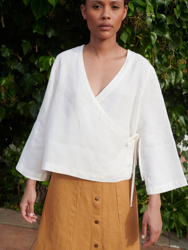 Model wearing a loose-fitting white linen top with a deep V-neck and long kimono sleeves paired with a camel linen skirt with metal snap buttons at the front