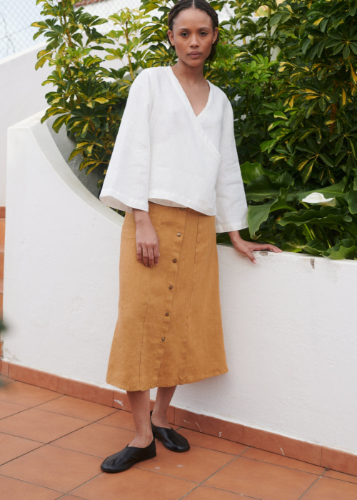 Model wearing a loose-fitting white linen V-neck wrap top and a camel color midi linen skirt with metal snap buttons