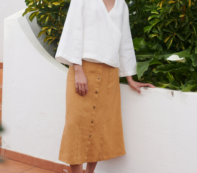 Model wearing a loose-fitting white linen V-neck wrap top and a camel color midi linen skirt with metal snap buttons
