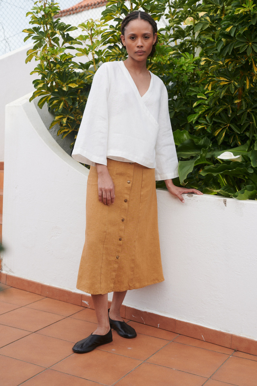 Model wearing a loose-fitting white linen V-neck wrap top and a camel color midi linen skirt with metal snap buttons