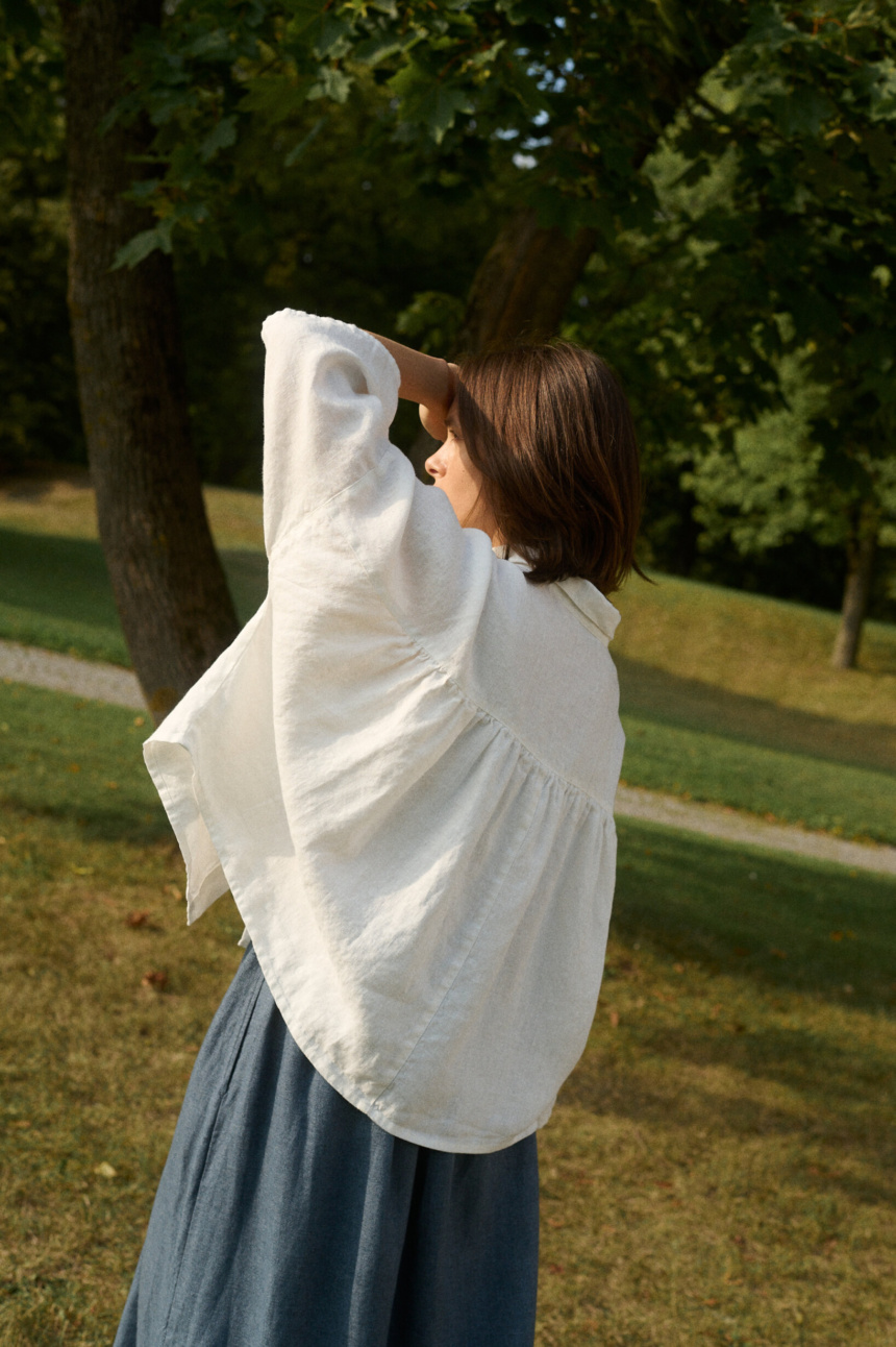 Model wearing a flowing white linen blouse with voluminous sleeves and a gathered back, paired with a dark blue linen wool blend skirt