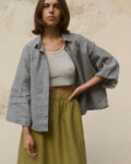 Woman wearing a light grey, loose-fitting linen button-up shirt with wide sleeves with a ribbed white cropped tank top underneath, paired with a high-waisted olive green heavy linen skirt featuring an elastic waistband