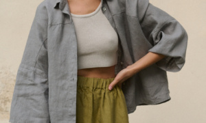 Woman wearing a light grey, loose-fitting linen button-up shirt with wide sleeves with a ribbed white cropped tank top underneath, paired with a high-waisted olive green heavy linen skirt featuring an elastic waistband