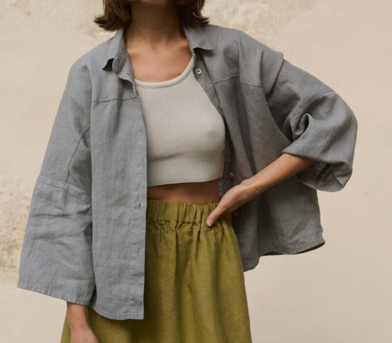 Woman wearing a light grey, loose-fitting linen button-up shirt with wide sleeves with a ribbed white cropped tank top underneath, paired with a high-waisted olive green heavy linen skirt featuring an elastic waistband