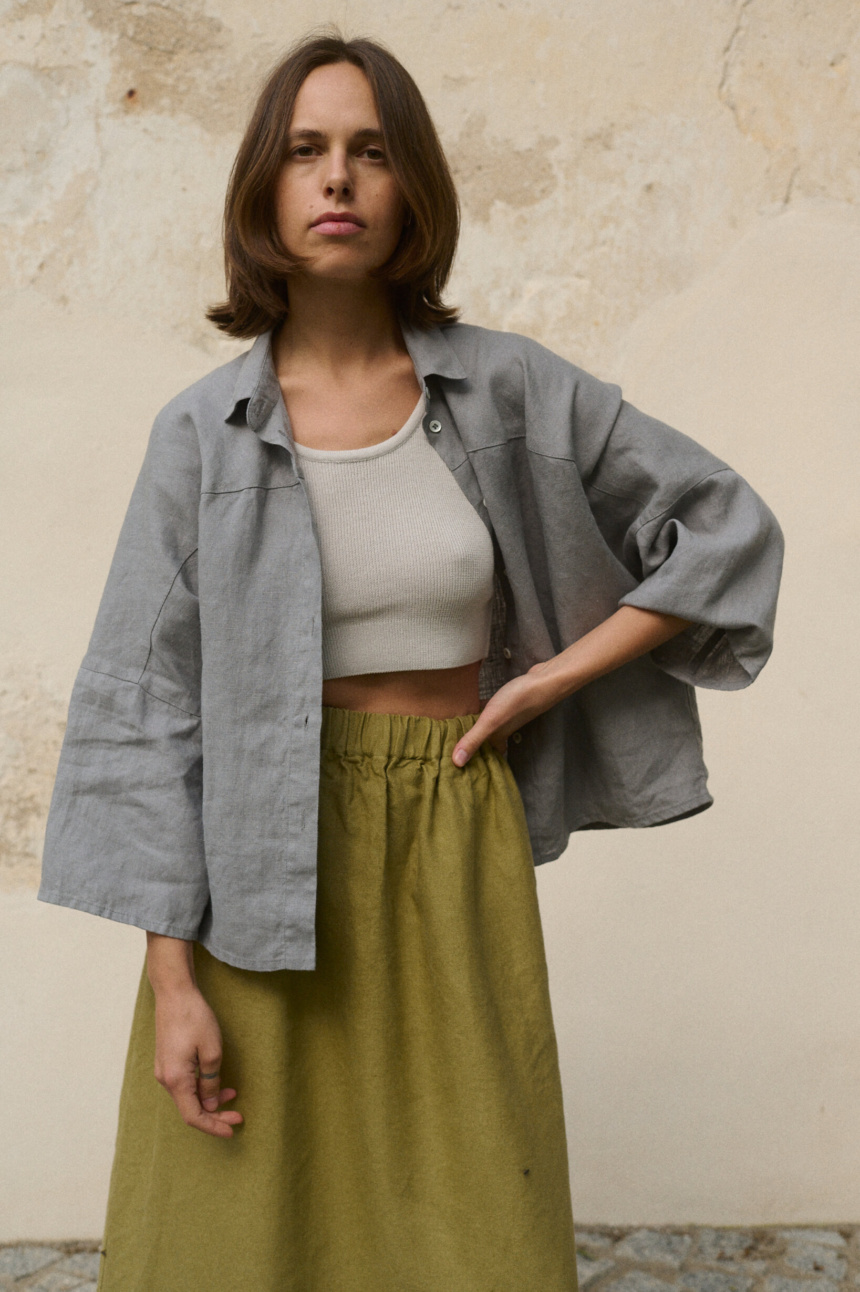 Woman wearing a light grey, loose-fitting linen button-up shirt with wide sleeves with a ribbed white cropped tank top underneath, paired with a high-waisted olive green heavy linen skirt featuring an elastic waistband