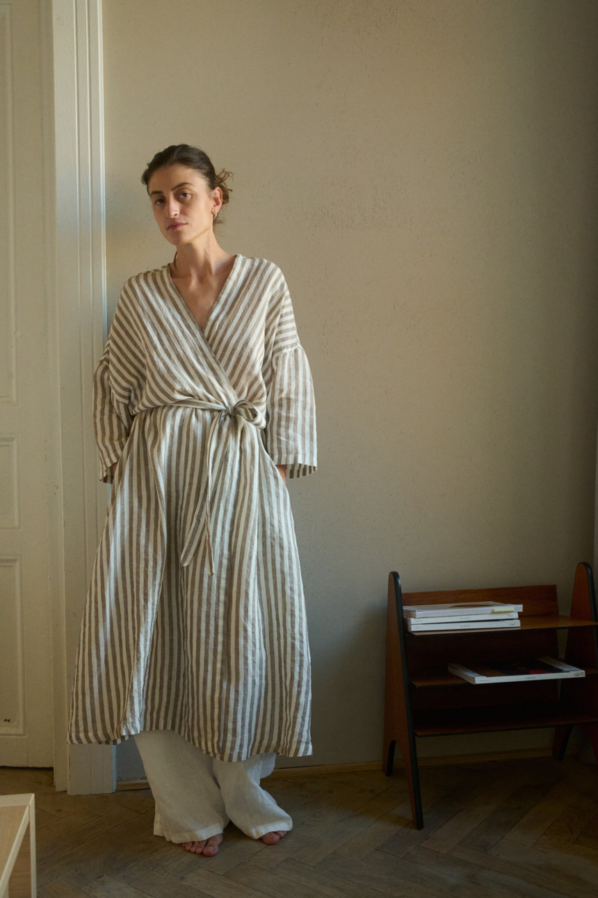 Woman wearing a long cacao stripes linen robe tied at the waist, layered over white pants, all made from lightweight linen